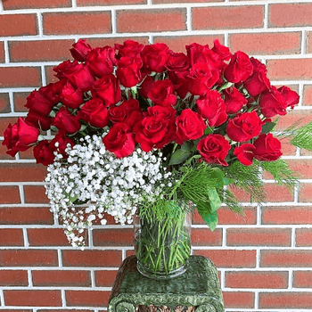 A glass vase of 50 red roses and baby's breath sitting on a stand.