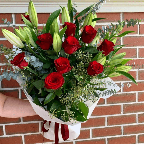 Red roses and lilies bouquet wrapped in white paper with burgundy ribbon.