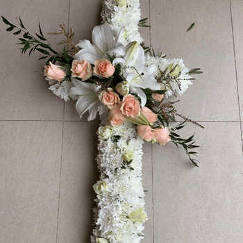Funeral cross with white and peach flowers sitting on the floor.